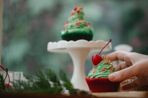 A hand reaching for a Christmas-themed cupcake with green frosting and cherry topping.